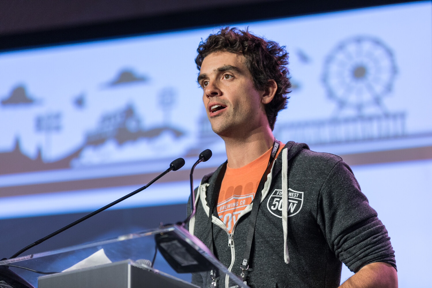 A man with dark curly hair in a gray Fifty West hoodie over an orange Brewing Co. T-shirt speaks at a clear podium in front of a slide with a Ferris wheel skyline illustration.