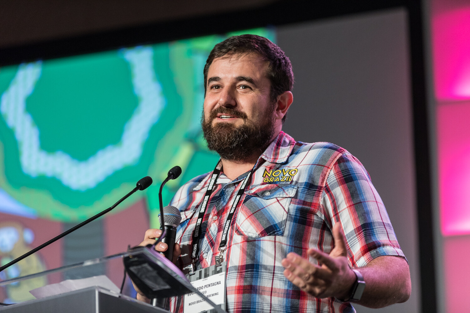 A bearded man in a red and blue plaid shirt with a Novo Brazil logo gestures with one hand at a clear podium in front of a green and white projected slide.