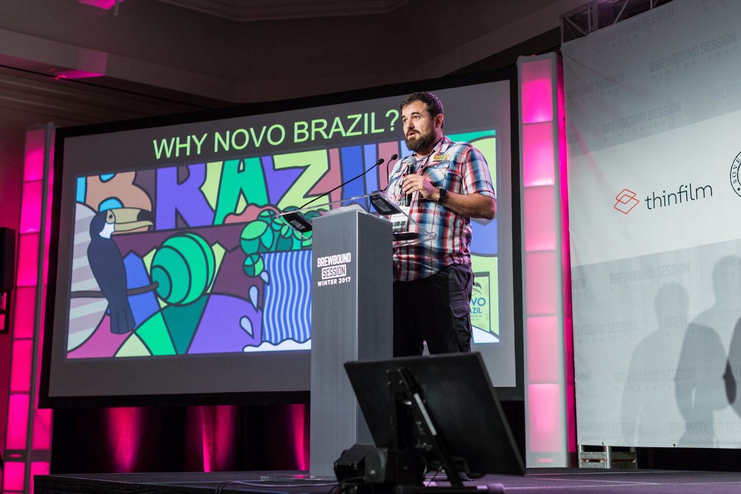 A bearded man in a plaid shirt speaks into a microphone at a podium on a stage beside a large screen reading Why Novo Brazil with a colorful toucan illustration.