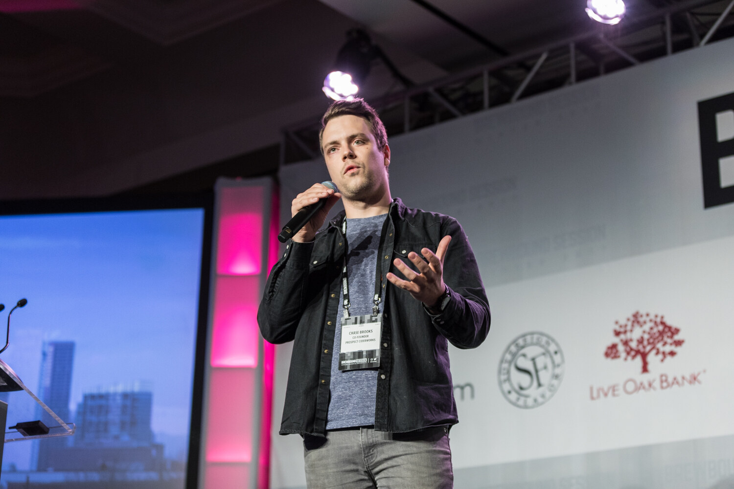 A man in a dark jacket over a gray T-shirt with a conference badge holds a microphone and gestures with one hand in front of Live Oak Bank and Sovereign Flavors logos and a screen showing a city skyline.