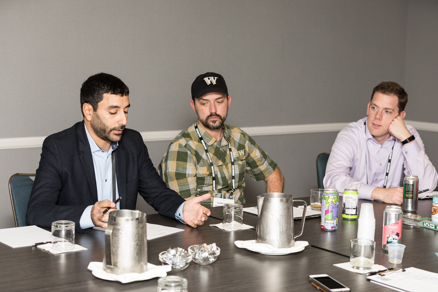 A man in a dark blazer gestures while a man in a black W cap and a man in a lavender shirt resting his chin on his hand listen at a long table with metal pitchers, beer cans and papers.