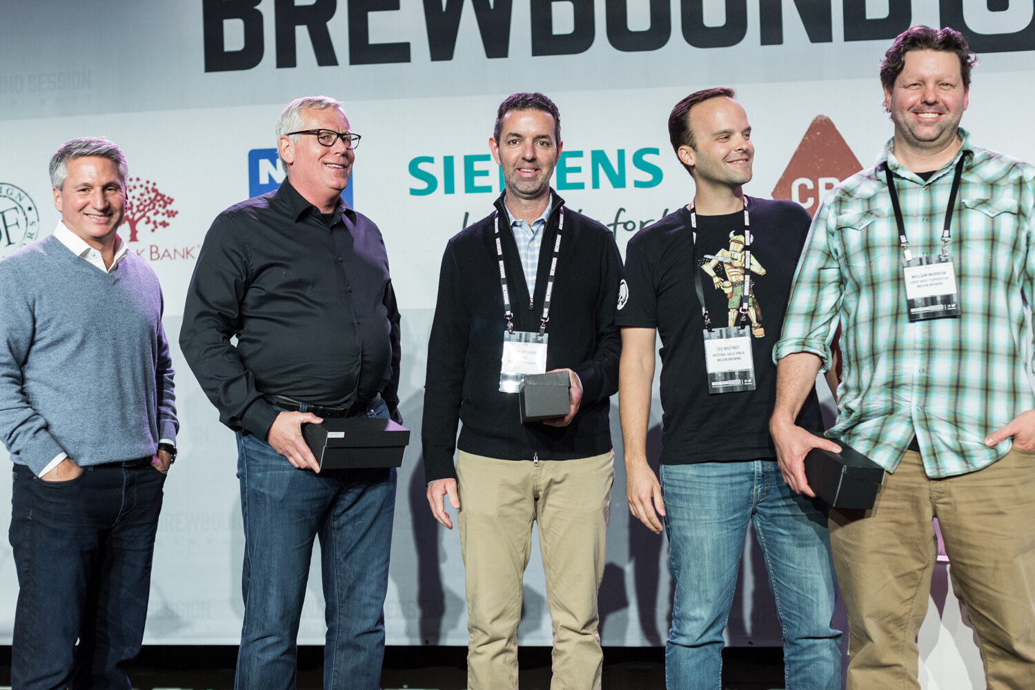 Five smiling men with conference badges stand in a row on a stage in front of a Brewbound Session backdrop with Siemens and Live Oak Bank logos, three of them holding small black boxes.