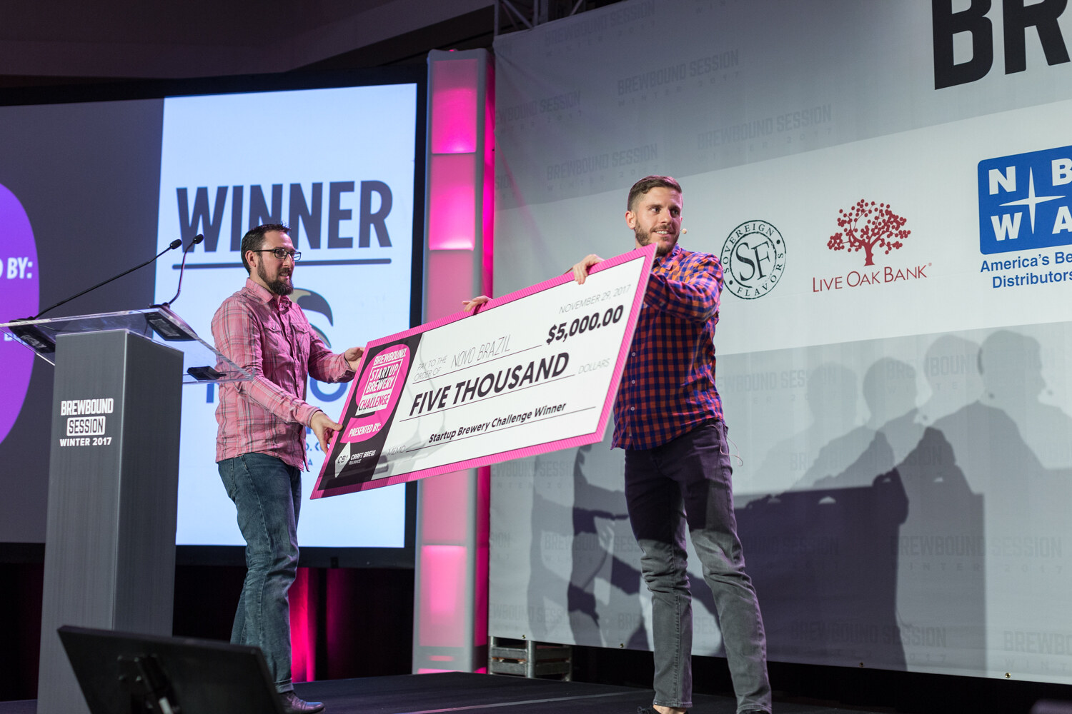 A man in a pink plaid shirt and a bearded man in a blue and orange checked shirt hold a large pink check reading Five Thousand dollars to the order of Novo Brazil beside a podium and a screen reading Winner.