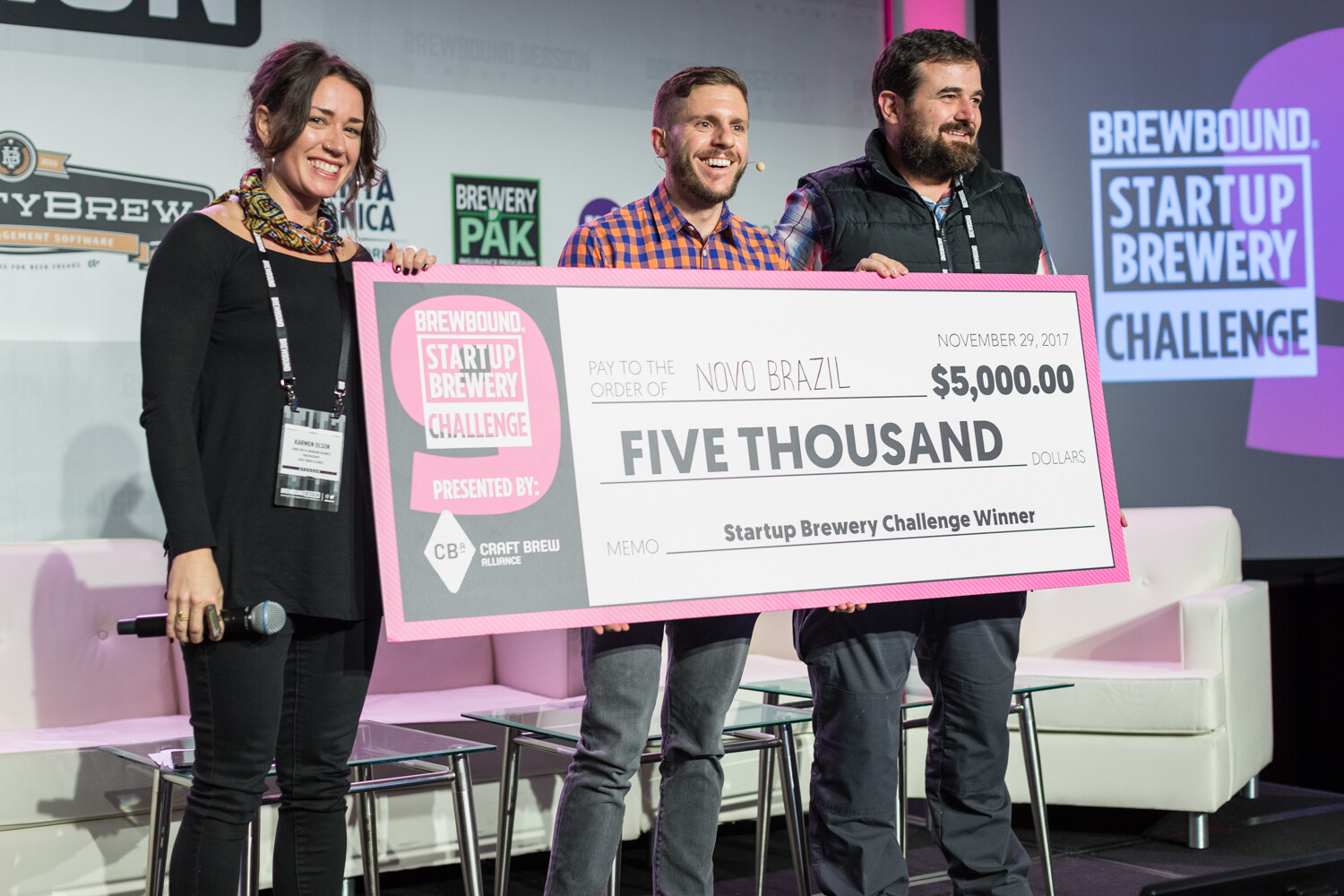 A woman in black holding a microphone and two men hold a large pink Brewbound Startup Brewery Challenge 9 check for five thousand dollars made out to Novo Brazil, with white sofas and a screen behind them.
