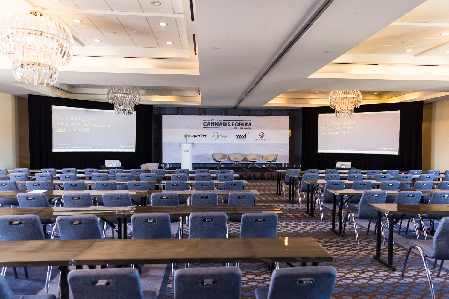 An empty hotel ballroom with long tables, blue chairs, chandeliers and a stage with a Cannabis Forum banner and screens.