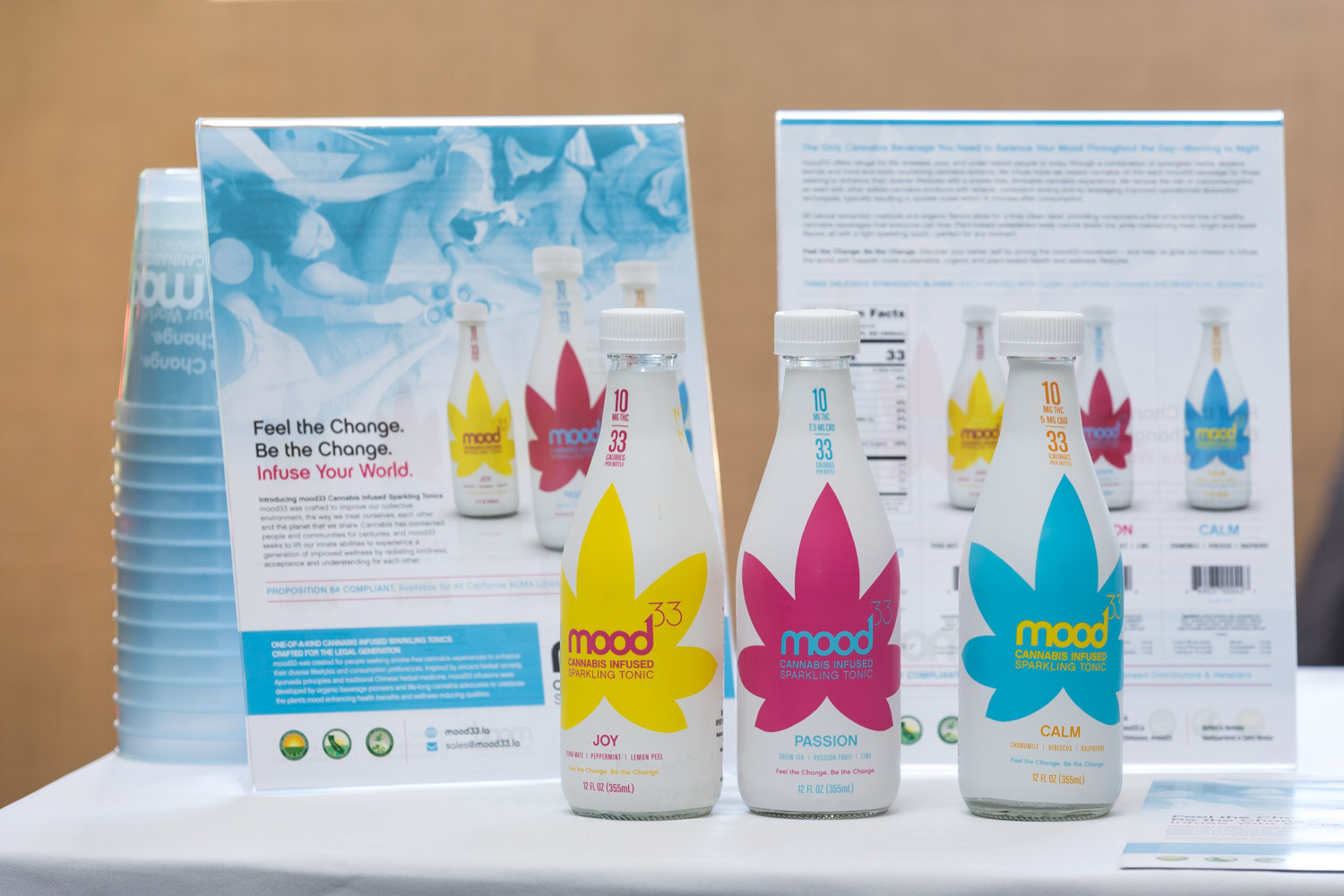 Three tall bottles with leaf-shaped labels in yellow, pink and blue beside a stack of cups and flyers.