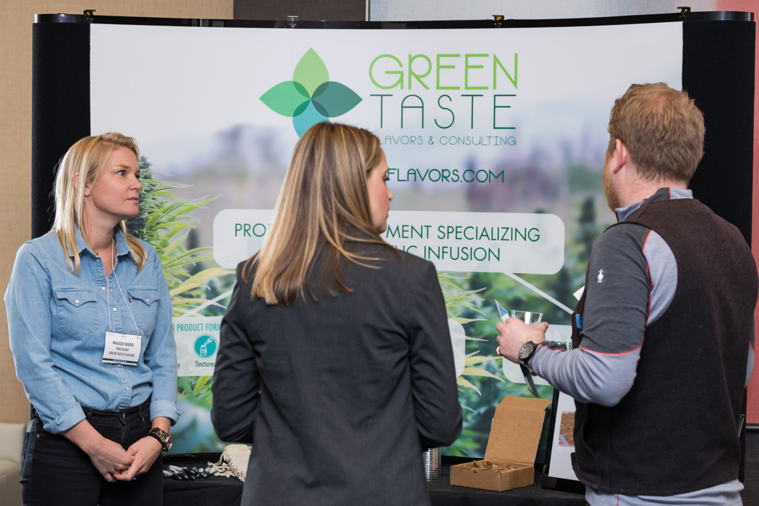 A woman in a denim shirt and two other attendees in front of a Green Taste Flavors and Consulting backdrop.