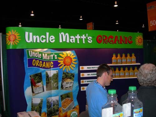 A booth with a green and purple Uncle Matt's Organic sign and orange juice bottles on shelves. A man in a blue shirt stands in front, with juice bottles in the foreground.