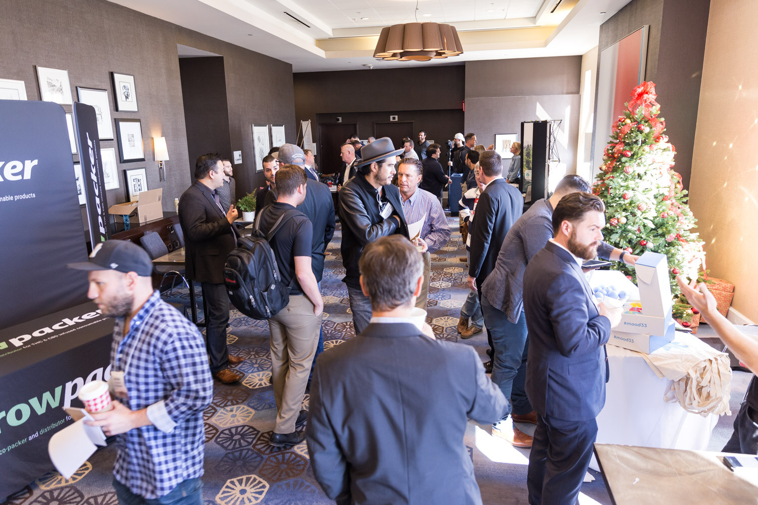 A crowded hotel hall with people talking beside a black growpacker display and a Christmas tree.