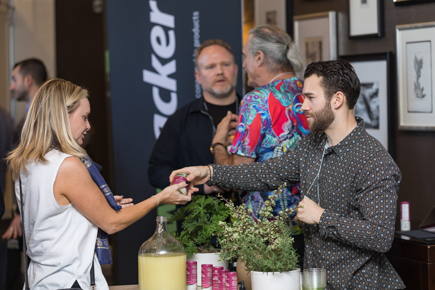 A woman in a white sleeveless top takes a small bottle from a man in a dark patterned shirt at a table with plants and a glass jar of juice.