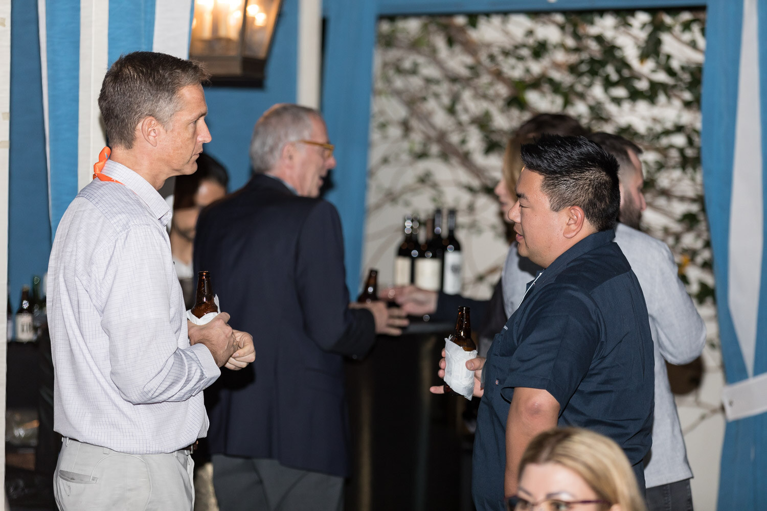 Several guests holding beer bottles talk in front of a blue striped cabana with flowers behind.