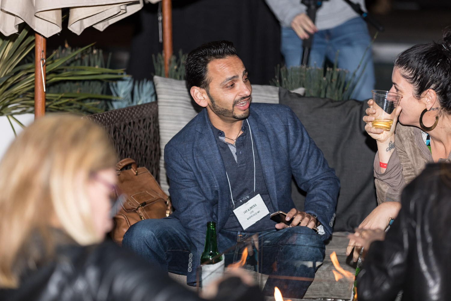 A man in a blue blazer with a name tag talks with a woman holding a drink beside a fire pit and patio umbrella.