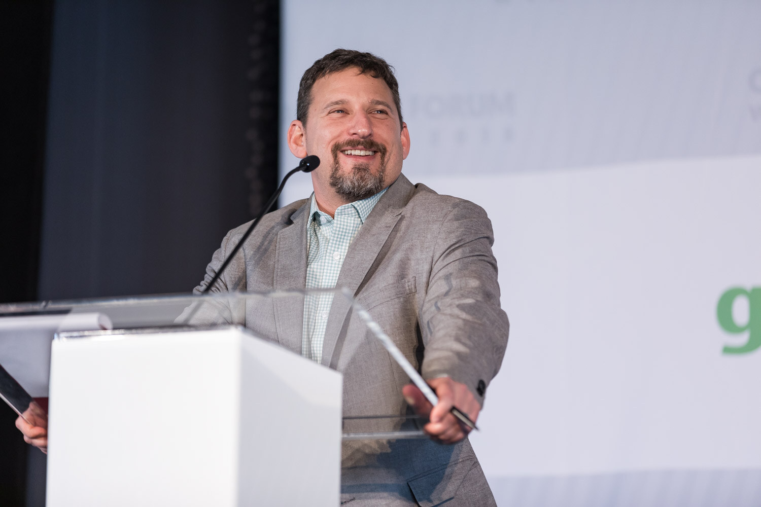 A man in a gray blazer smiles at a white lectern with a clear acrylic top in front of a banner.