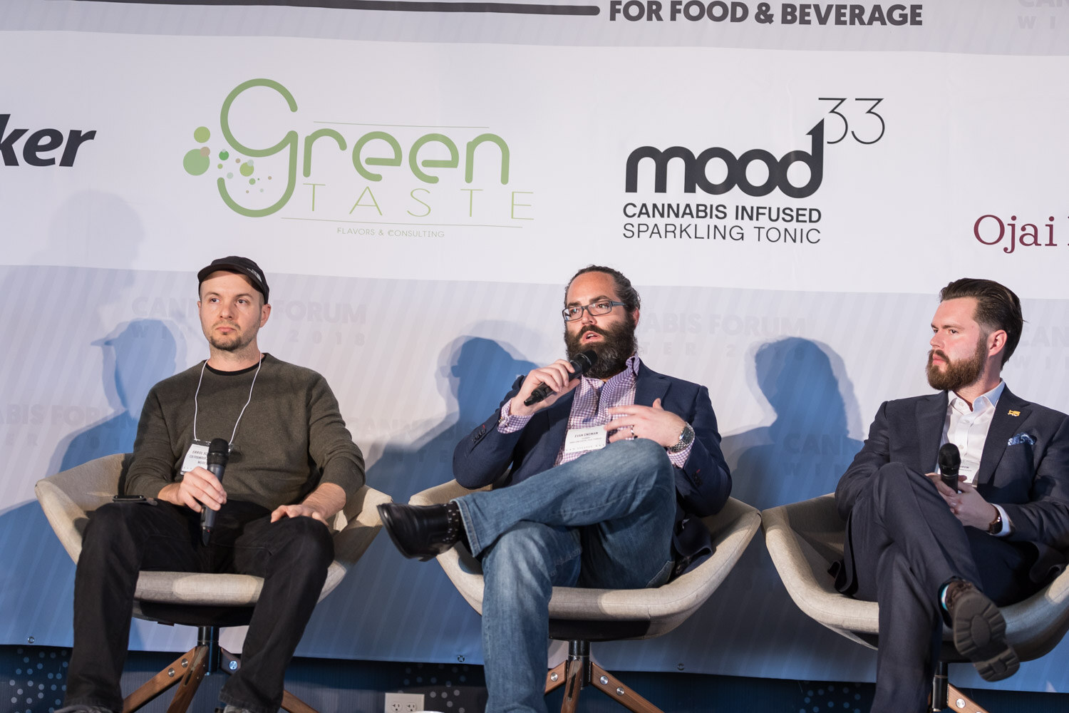 Three men in swivel chairs, one holding a microphone, in front of Green Taste and mood33 logos.