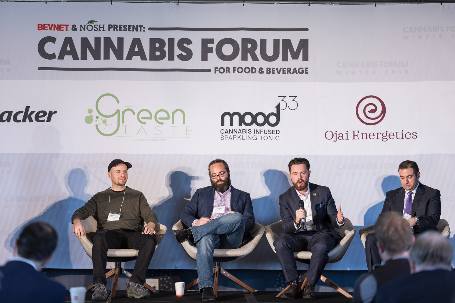 Four seated men on stage in front of a Cannabis Forum backdrop, with the heads of audience members at the bottom of the frame.