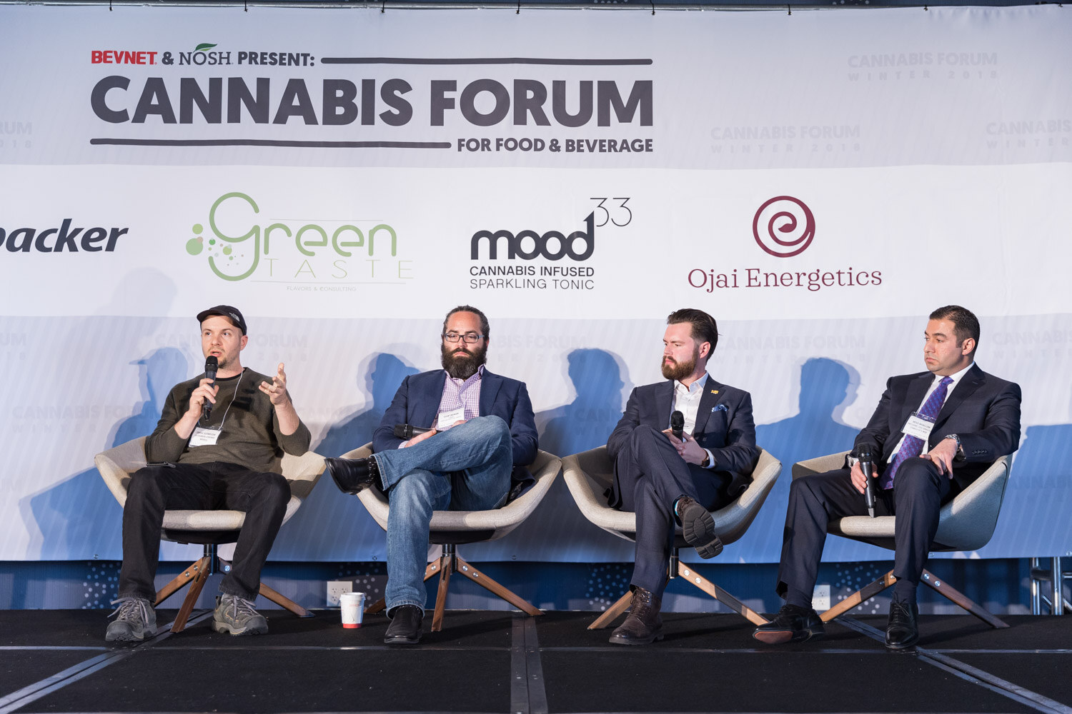Four men on white swivel chairs on a stage in front of a Cannabis Forum for Food and Beverage banner with sponsor logos.