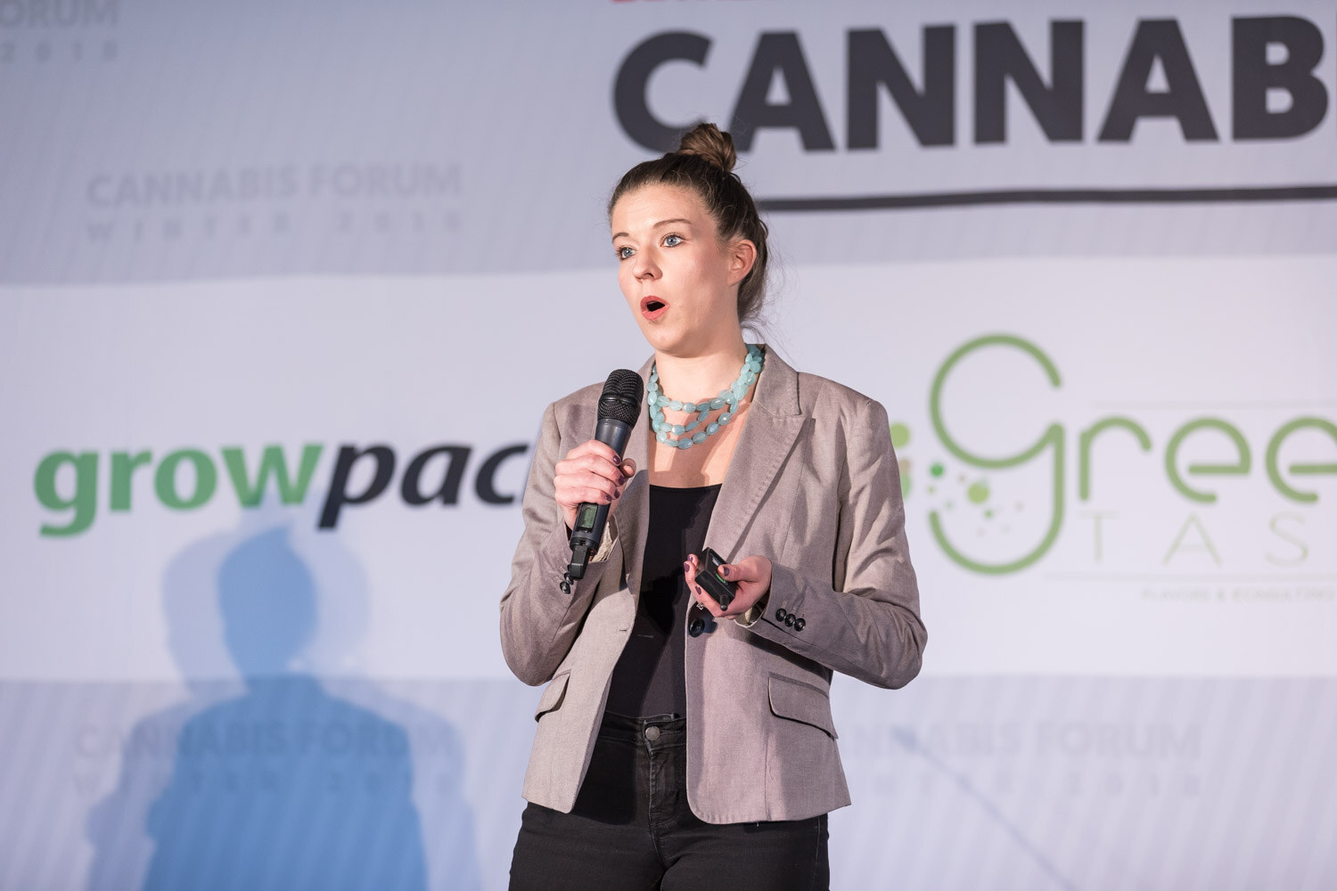 A woman in a gray blazer with her hair in a bun speaks into a microphone in front of a growpacker and Green Taste backdrop.