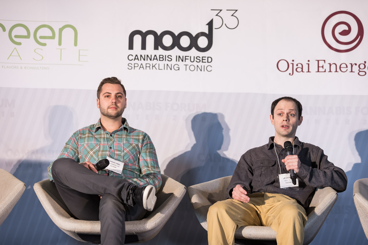 Two men in white swivel chairs, one in a green plaid shirt and one in a dark button-up with khaki pants, in front of mood33 and Ojai Energetics logos.