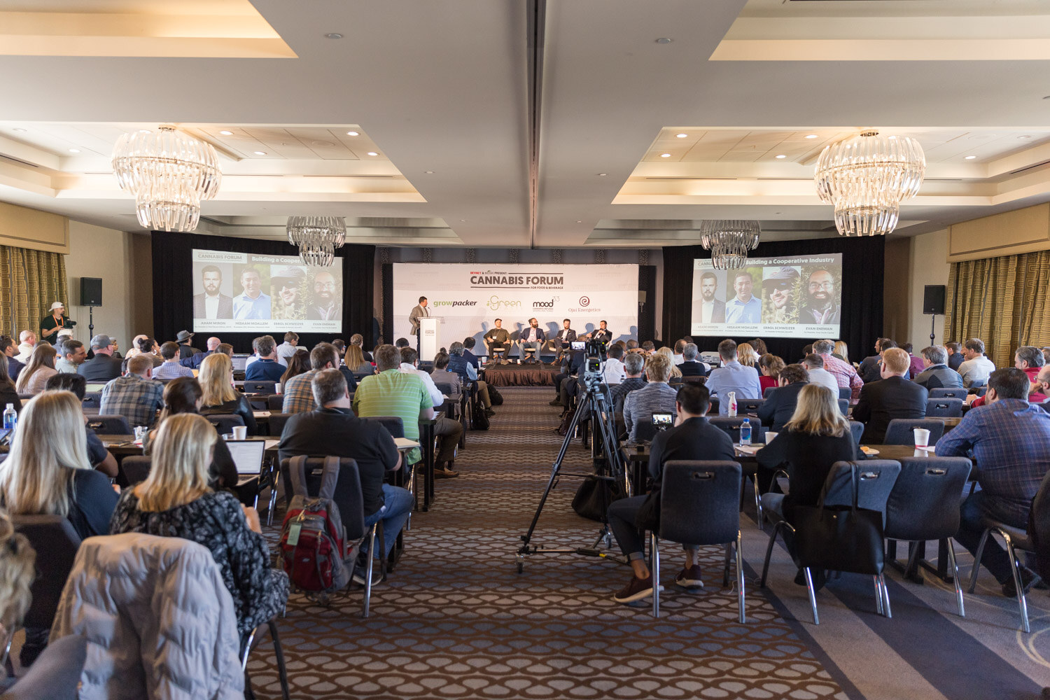 A wide view of a hotel ballroom with rows of attendees facing a stage and two large screens.