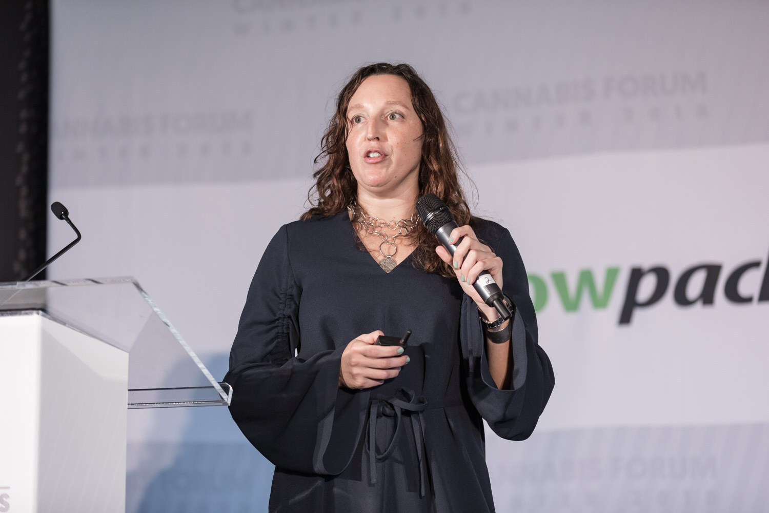 A woman with wavy brown hair in a dark dress speaks beside a clear lectern in front of a growpacker banner.
