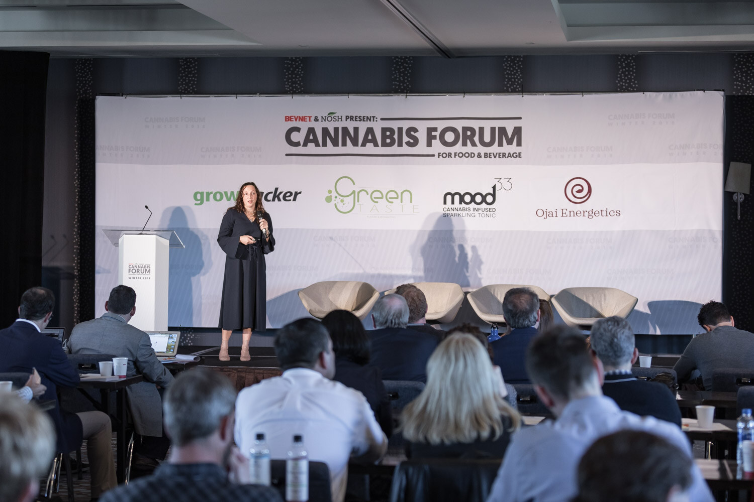A woman standing on a stage beside a lectern in front of the Cannabis Forum banner, with audience members at tables in the foreground.