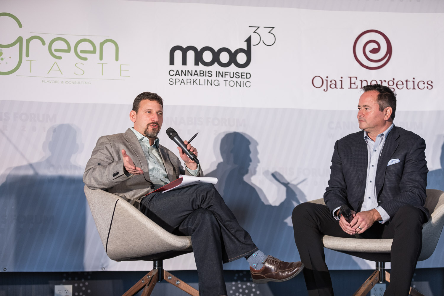Two men in beige swivel chairs, one holding a microphone and notes, in front of Green Taste, mood33 and Ojai Energetics logos.