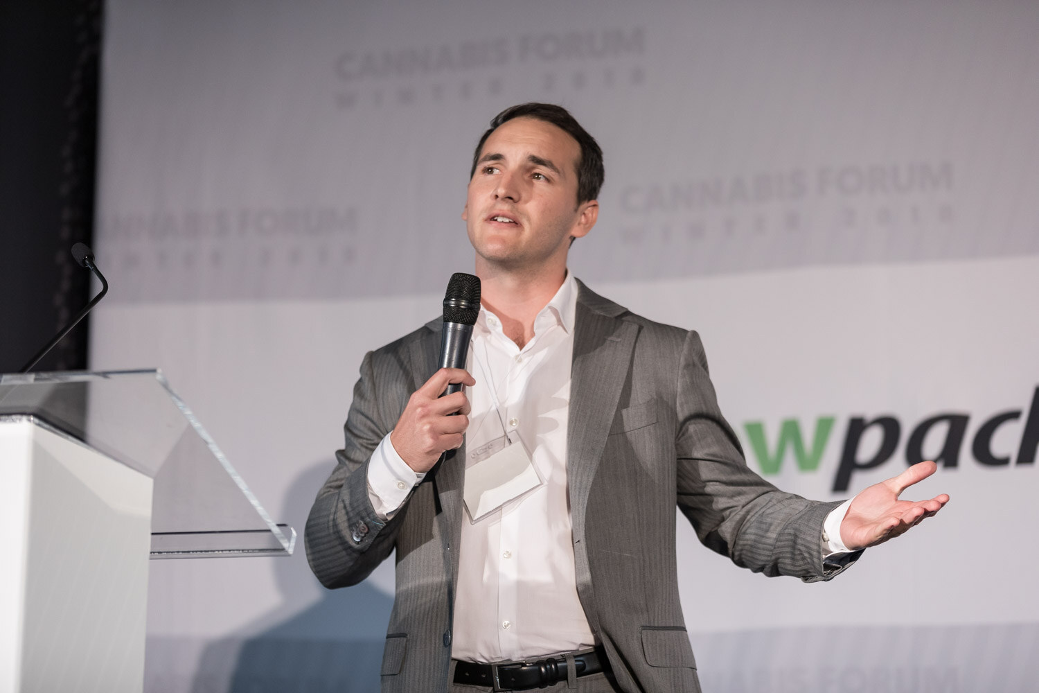 A man in a gray suit and open-collar white shirt speaks into a microphone with a clear lectern at left.