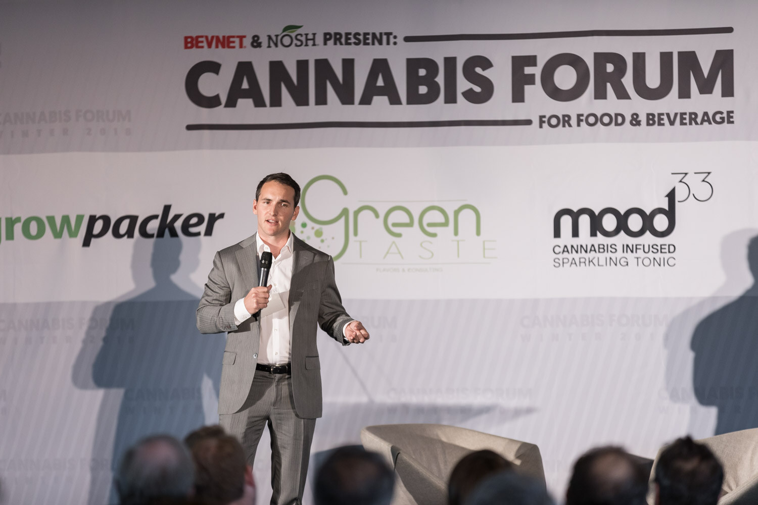 A man in a gray suit stands on stage with a microphone below a Cannabis Forum banner, audience heads in the foreground.