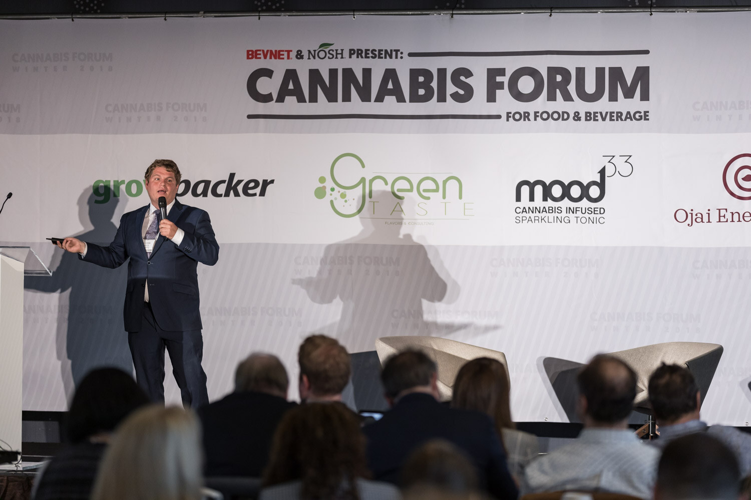 A man in a dark suit stands on stage in front of a Cannabis Forum banner with sponsor logos, with the audience in the foreground.