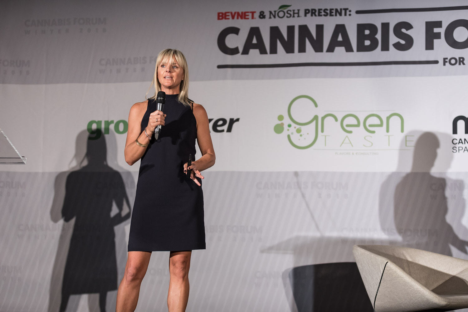 A woman with blond hair in a black sleeveless dress holds a microphone in front of a Cannabis Forum backdrop.