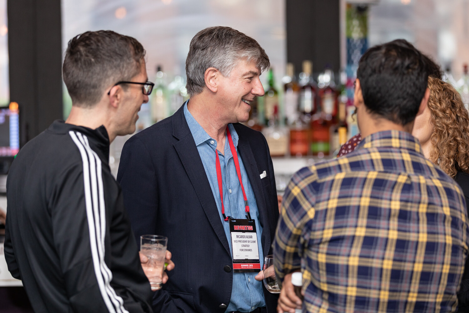 Three men stand in conversation holding drinks in front of a back bar of liquor bottles, one in a black track jacket and one in a navy blazer wearing a lanyard.