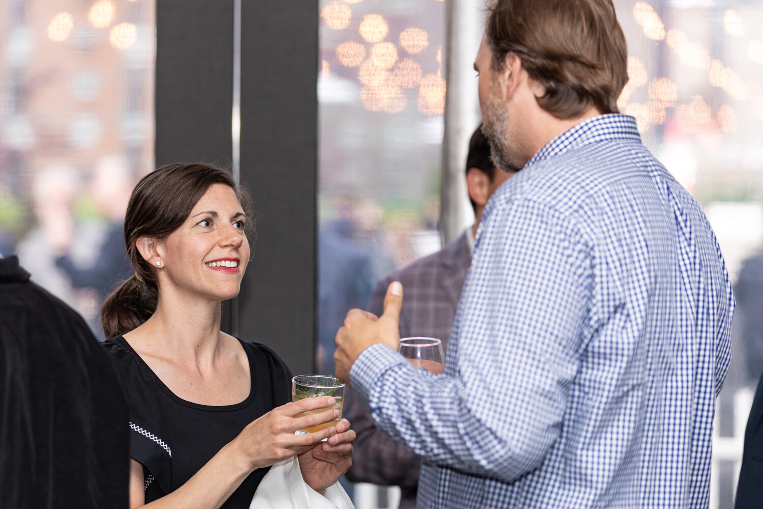 A woman in a black top with her hair pulled back holds a glass and smiles toward a tall man in a blue gingham shirt, with blurred city lights behind them.