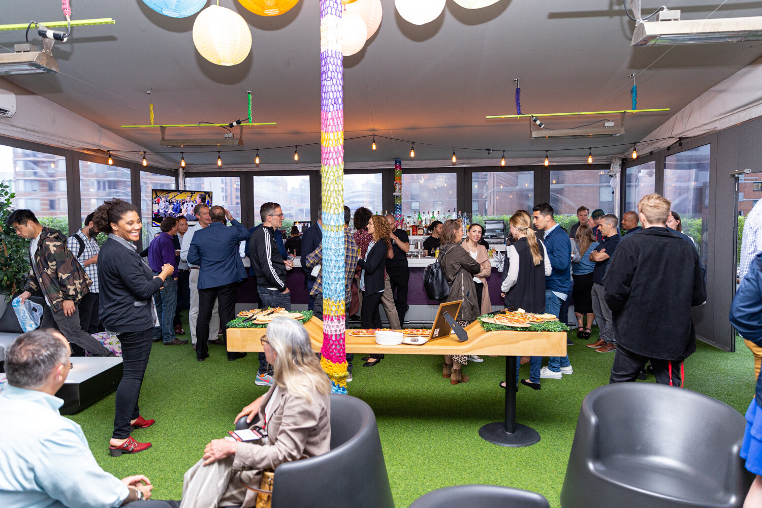 A crowd of guests stands on artificial turf around a wooden food table in a glass-walled room with colorful paper lanterns hanging from the ceiling.