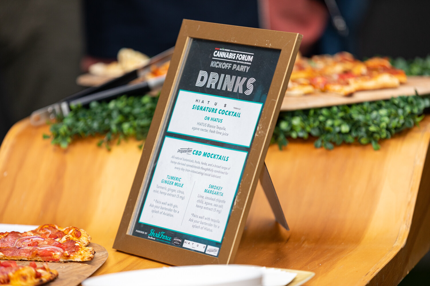 A gold-framed card headed Cannabis Forum Kickoff Party Drinks lists a signature cocktail and CBD mocktails, set on a wooden counter beside pizza slices and greenery.