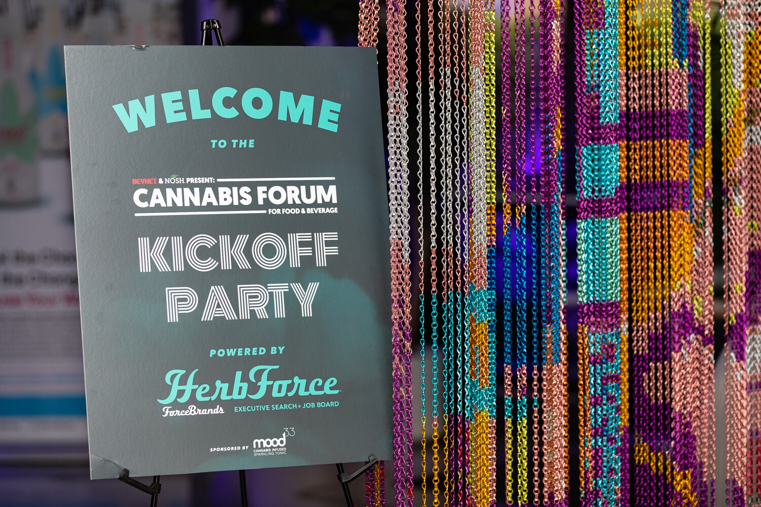 A dark easel sign reading Welcome to the BevNET & Nosh Present: Cannabis Forum Kickoff Party, Powered by HerbForce stands beside a colorful beaded chain curtain.