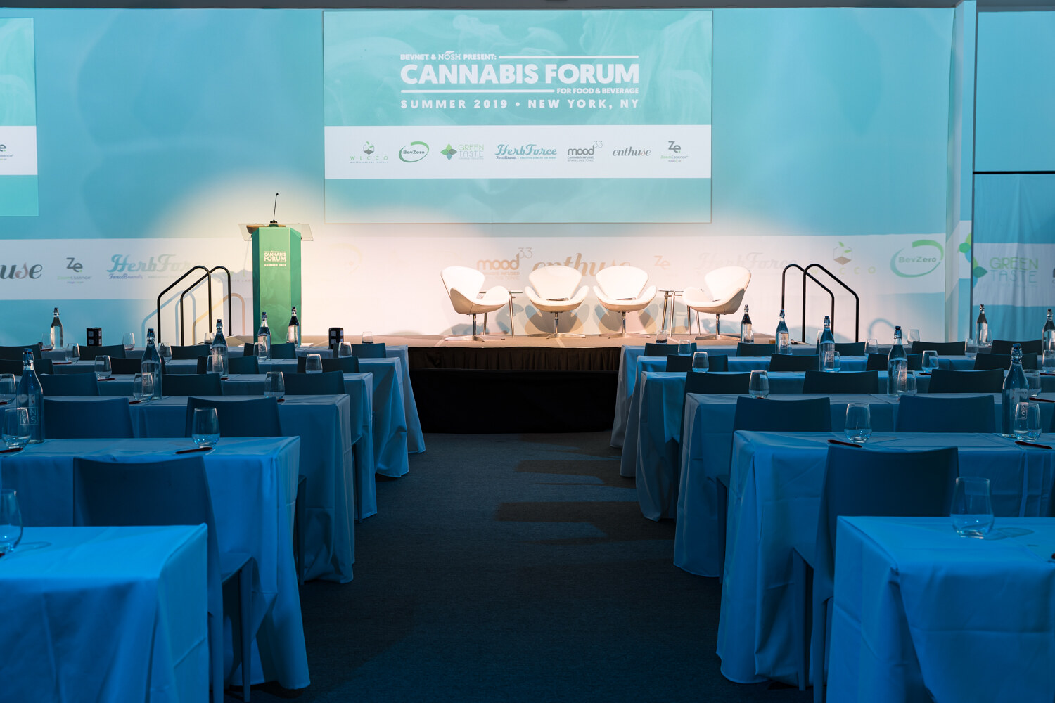 An empty room of blue-draped tables faces a lit stage with white lounge chairs and a screen reading Cannabis Forum for Food & Beverage, Summer 2019, New York, NY.