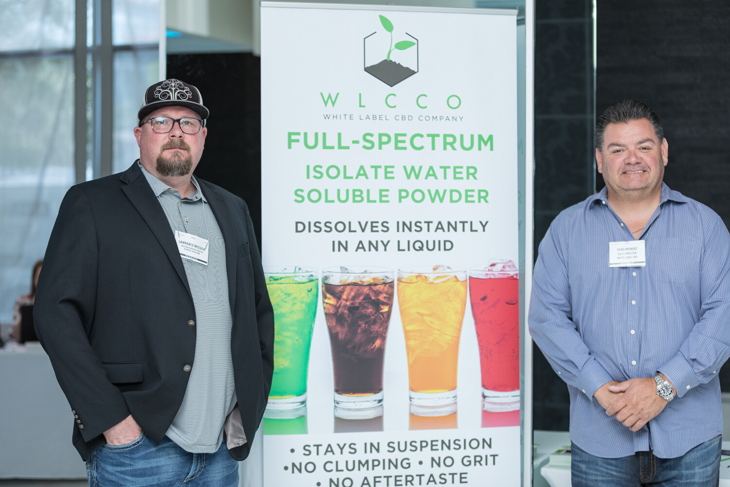 Two men with name badges stand beside a tall banner for White Label CBD Company advertising full-spectrum isolate water soluble powder with four glasses of colored drinks.