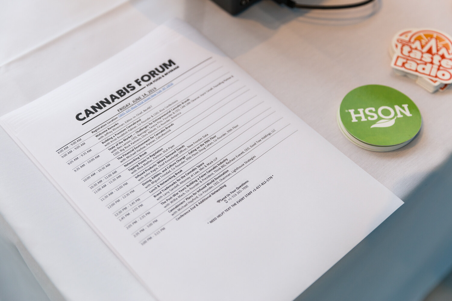 A sheet of paper headed Cannabis Forum with a timed agenda lies on a white tablecloth next to a round green HSON sticker and a partly visible Taste Radio sticker.