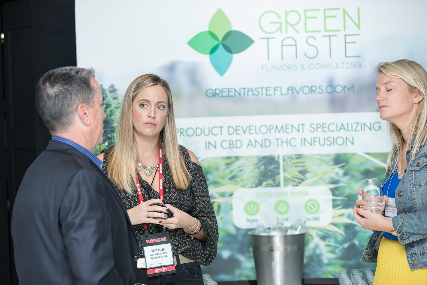A woman with a lanyard talks with a man and another woman in front of a Green Taste banner that reads Product development specializing in CBD and THC infusion.