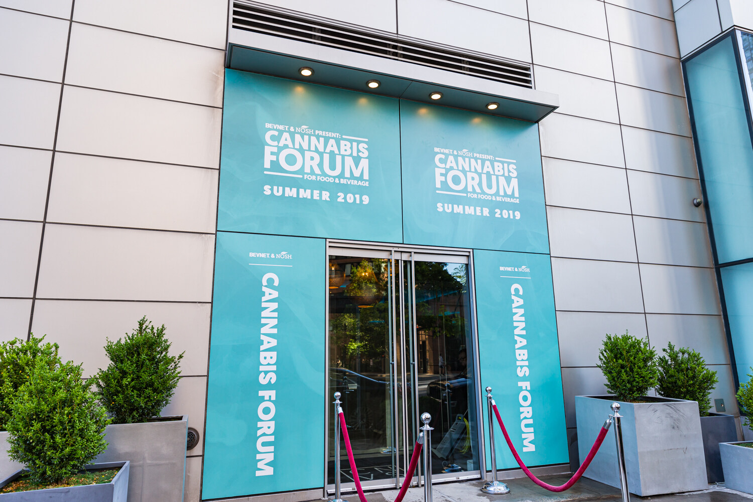 The glass entrance of a building is covered in teal panels reading BevNET & Nosh Present Cannabis Forum, Summer 2019, with red velvet ropes and potted shrubs at the door.