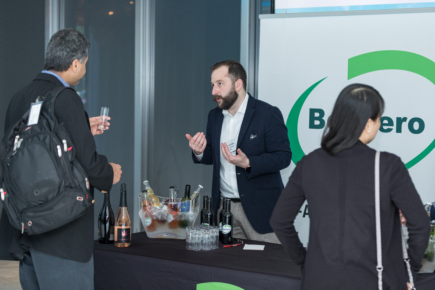 A bearded man in a navy blazer gestures behind a dark table of sparkling bottles and cups, with attendees in front and a partly visible BevZero banner behind him.