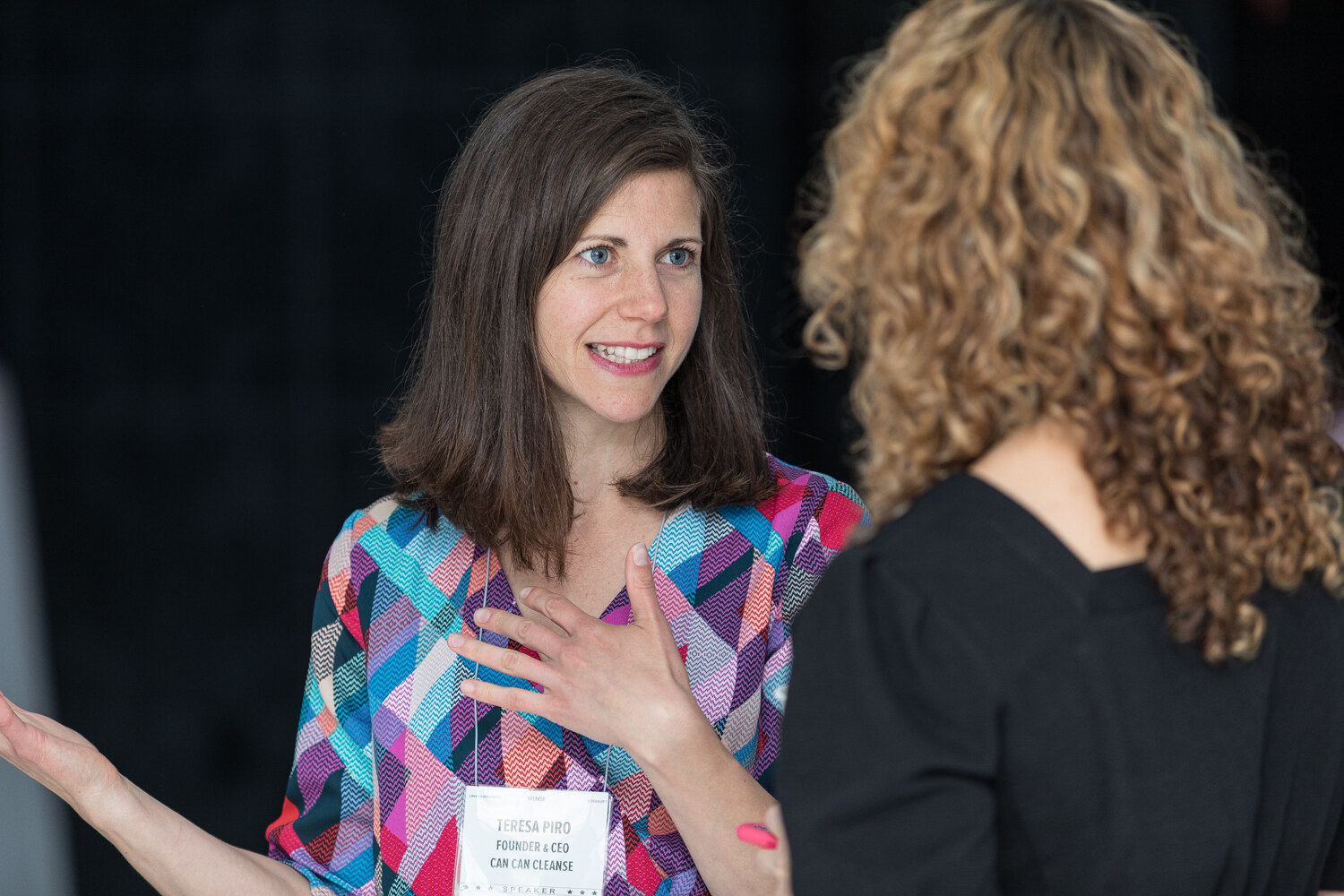 A woman in a colorful geometric-print dress with a name badge talks with a woman with curly hair, whose back is partly to the camera, against a dark wall.