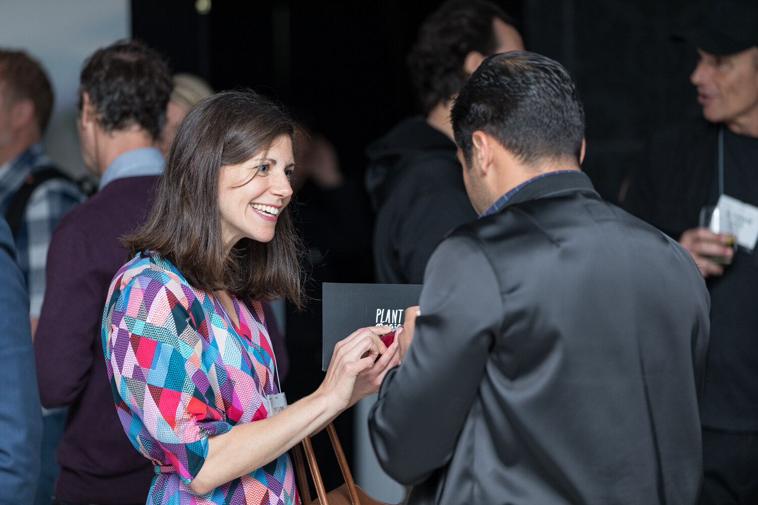 A smiling woman in a geometric-print dress holds a small black card toward a man in a dark jacket seen from behind in a crowded room.