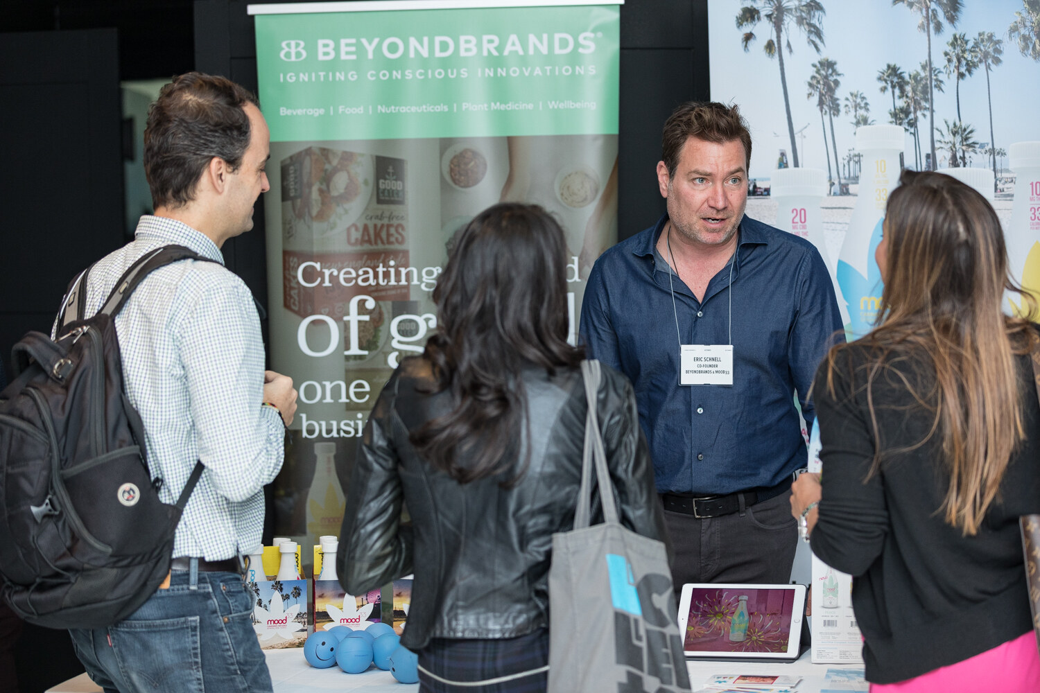 A man in a navy shirt with a name badge talks with attendees in front of a Beyond Brands banner and bottles of Mood33 beverages on the table.