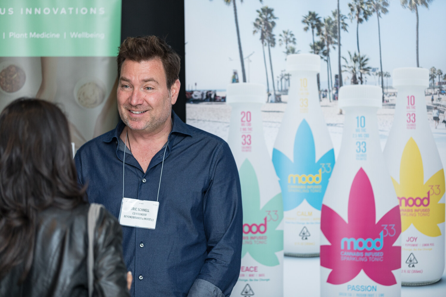 A man in a navy shirt with a name badge smiles in front of tall bottles labeled mood33 Cannabis Infused Sparkling Tonic in blue, pink and yellow leaf designs.