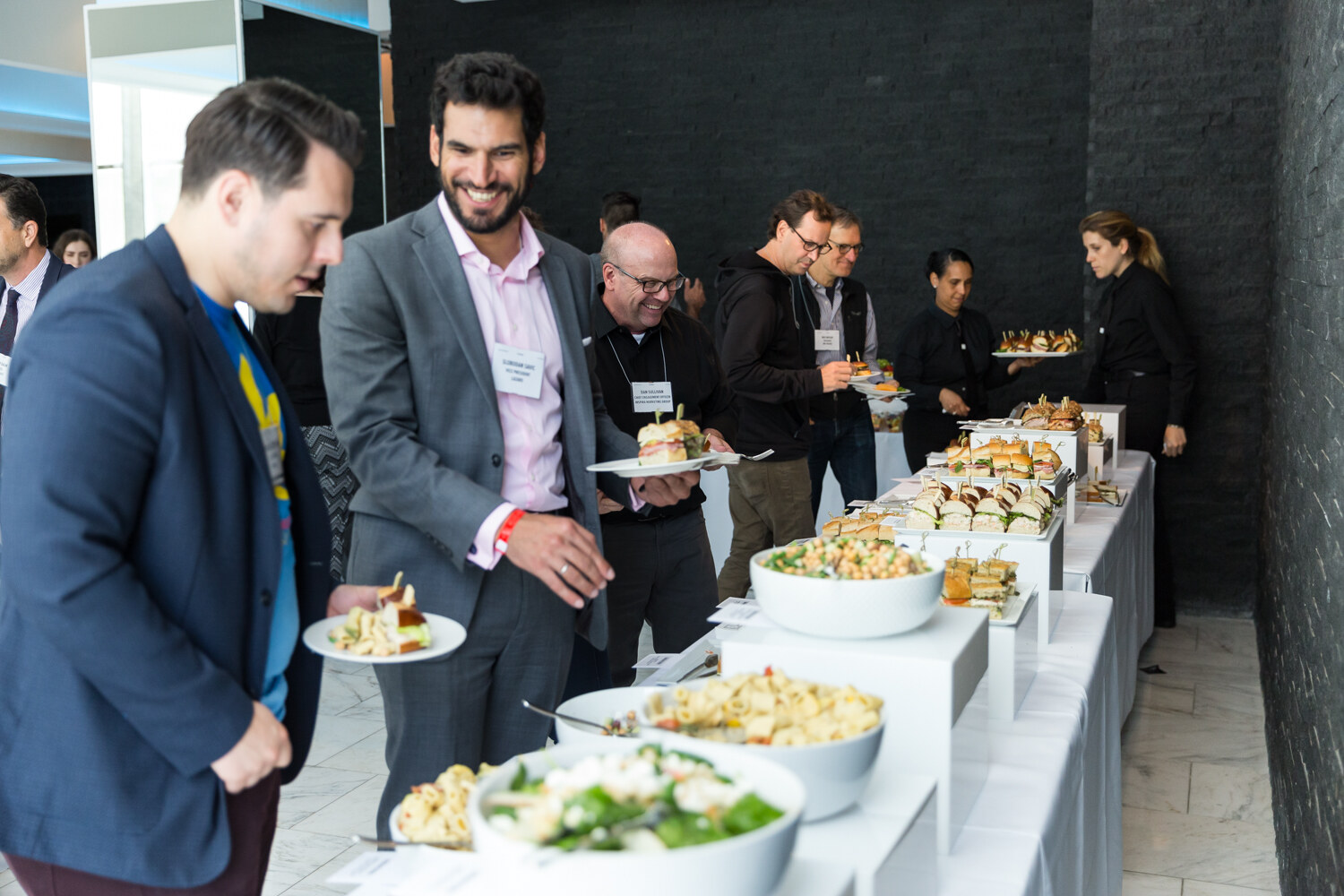Attendees with plates stand along a long buffet of white platforms holding bowls of salad and platters of sandwiches against a dark stone wall.