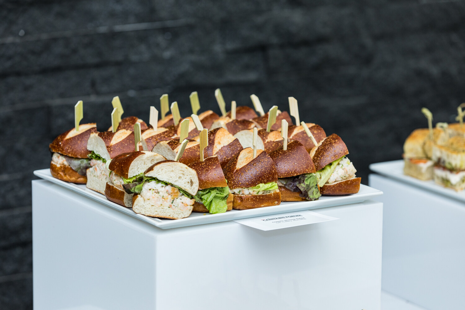 A white platter of small sandwiches on pretzel rolls with lettuce and a filling, each topped with a pick, on a white pedestal.