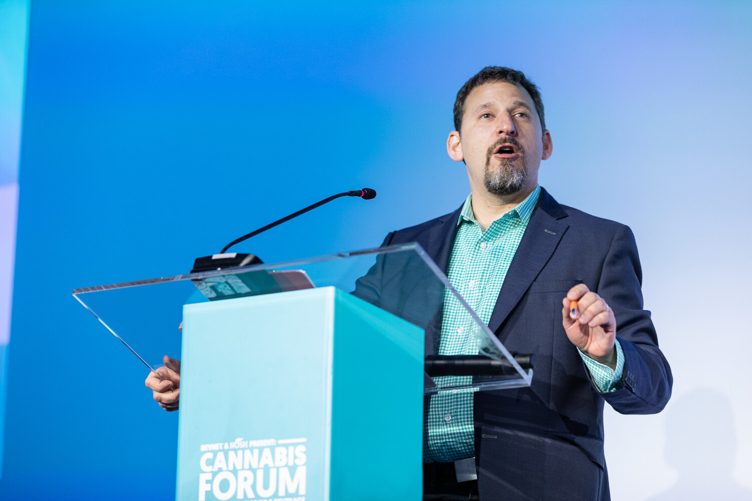 A bearded man in a navy blazer and teal checked shirt speaks into a microphone on a clear podium against a bright blue background.