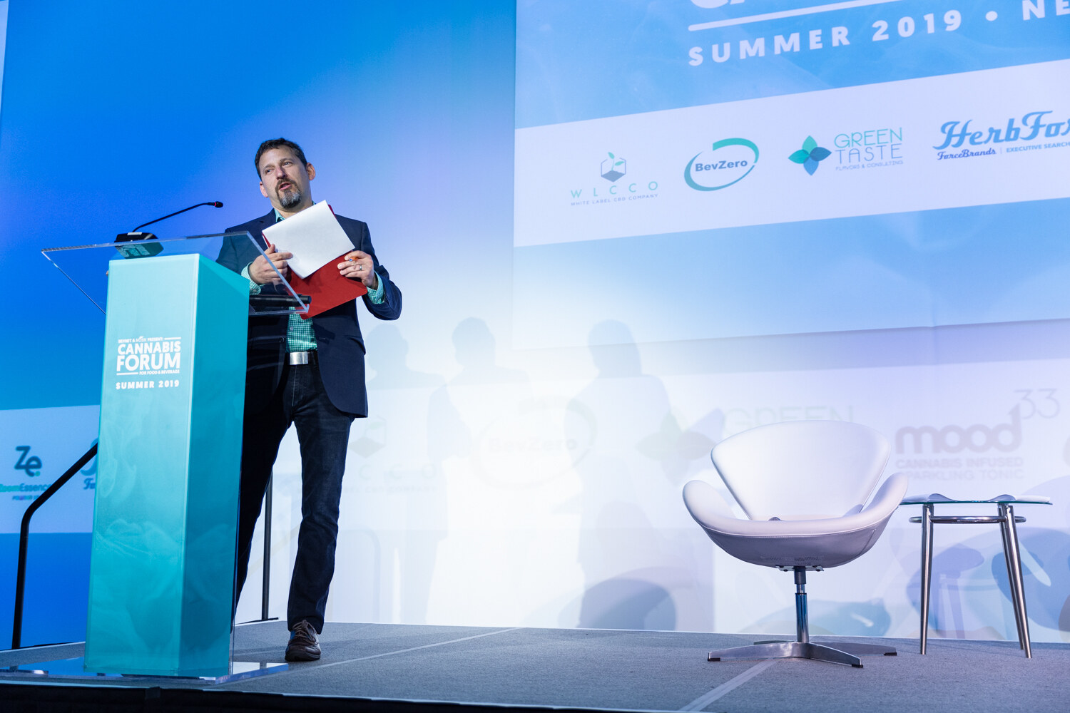 A bearded man in a navy blazer holds papers at a teal Cannabis Forum podium on a bright blue stage, with an empty white lounge chair and sponsor logos behind him.