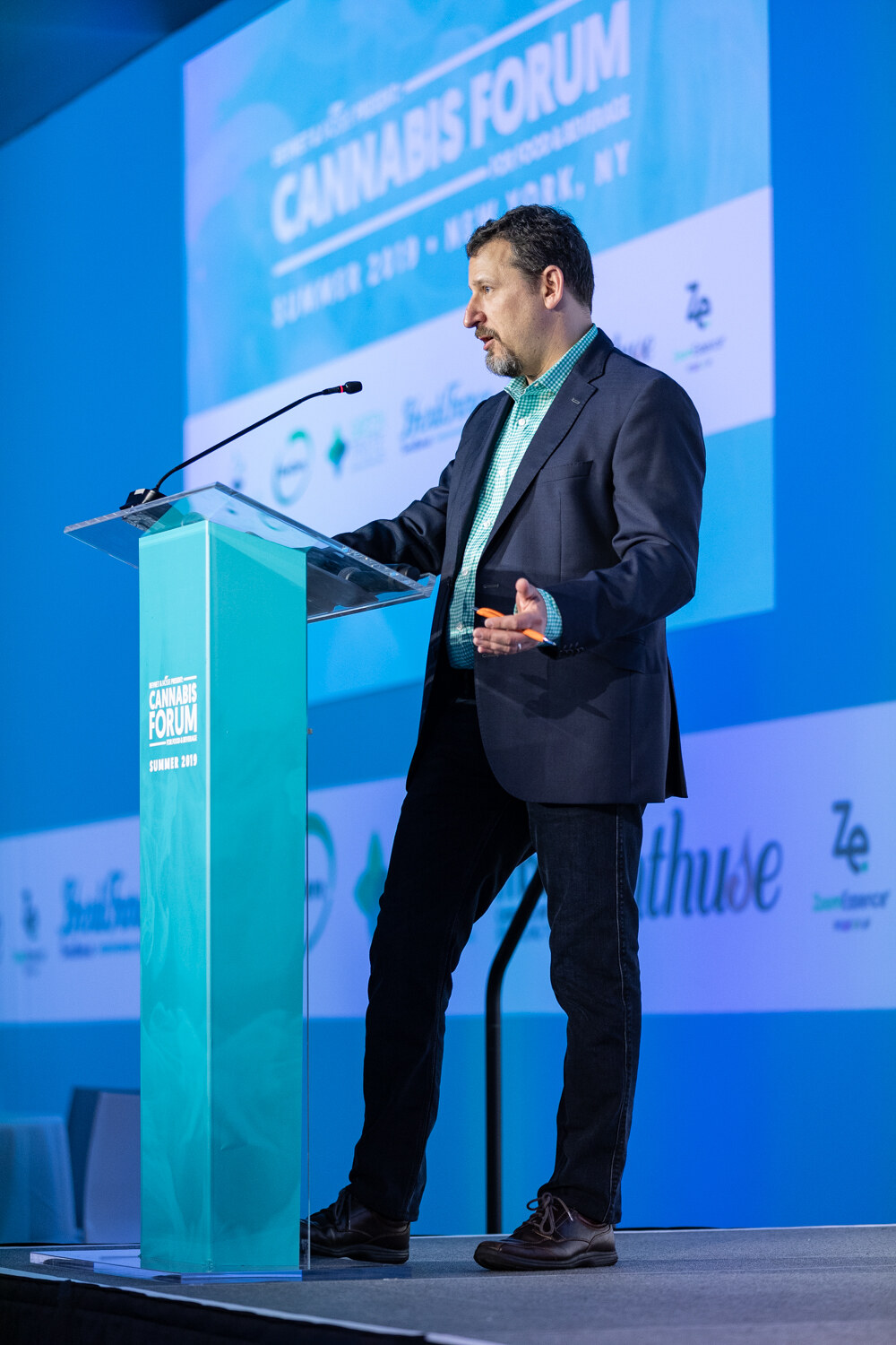 A man in a dark blazer gestures at a teal podium on a blue-lit stage, with a screen above reading Cannabis Forum Summer 2019 New York, NY.